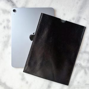 Può includere: Un tablet argentato con un logo Apple nero è accanto a una custodia in pelle nera. La custodia ha un design semplice con un piccolo ritaglio nella parte superiore. Il tablet ha un obiettivo della fotocamera sul bordo superiore.