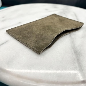 Può includere: Un piccolo porta carte in pelle scamosciata verde oliva. La custodia rettangolare ha un'apertura curva ed è cucita lungo i bordi. Il materiale morbido e strutturato è visibile in questa ripresa ravvicinata. Il porta carte è progettato per proteggere e trasportare le carte.