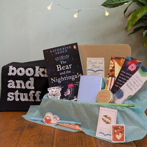 Puede incluir: Una caja de regalo con libros y artículos de papelería. Incluye un ejemplar de "The Bear and the Nightingale", una bolsa de mano negra con el texto "books and stuff", marcadores, un cuaderno, pegatinas, bolígrafos y un lápiz. La caja está sobre papel de seda azul.