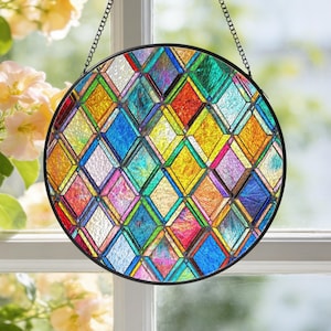 May include: A circular stained glass window suspended by a chain. The window displays a vibrant geometric diamond pattern in red, yellow, blue, green, and purple. It's framed in black and hangs in front of a window.