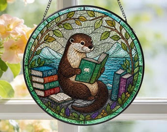 May include: A circular stained glass suncatcher featuring a brown otter reading a green book. The otter is surrounded by a stack of colorful books, a blue lake, and distant mountains, all framed by green leafy branches and a black metal chain.