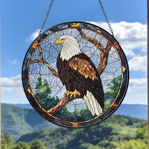 Puede incluir: Una vidriera redonda con un águila calva posada en una rama. El águila tiene plumas blancas y marrones, un pico amarillo, sobre un cielo azul y un paisaje verde. La ventana tiene un borde decorativo y cuelga de una cadena.