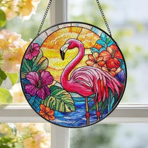 Puede incluir: Un atrapasol circular de vidrieras que muestra un flamenco rosa de pie en agua azul. El fondo presenta un sol amarillo, hojas tropicales verdes y vibrantes flores de hibisco moradas y naranjas, todo en un marco de metal negro.
