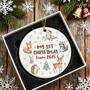 May include: A white ceramic Christmas ornament with a gold string, featuring woodland animals and the text "MY 1ST CHRISTMAS Liam 2025". The ornament is in a white box with a black interior, perfect for holiday gifting.