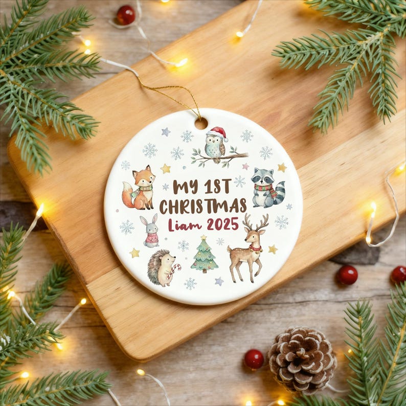 May include: A white ceramic Christmas ornament with a gold string, featuring woodland animals and the text "MY 1ST CHRISTMAS Liam 2025". The ornament has a festive design with snowflakes and stars, perfect for holiday decor.