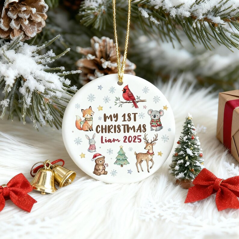 May include: A white ceramic Christmas ornament with a gold string. The ornament features the text "My 1st Christmas Liam 2025" and illustrations of a fox, koala, deer, and other festive animals. Snowflakes and stars decorate the design.