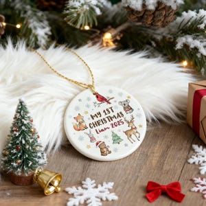 May include: A round Christmas ornament with a gold string, featuring the text "My 1st Christmas Liam 2025" and illustrations of animals. The ornament is white with a gold string, and is surrounded by Christmas decorations.