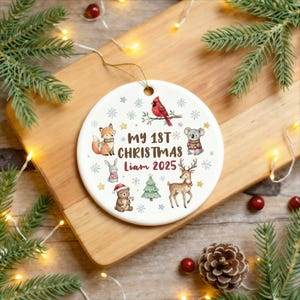 May include: A white ceramic Christmas ornament with a gold string. The ornament features a red cardinal, a fox, a koala, a deer, a bear, a rabbit, and a Christmas tree. Text reads "My 1st Christmas Liam 2025".