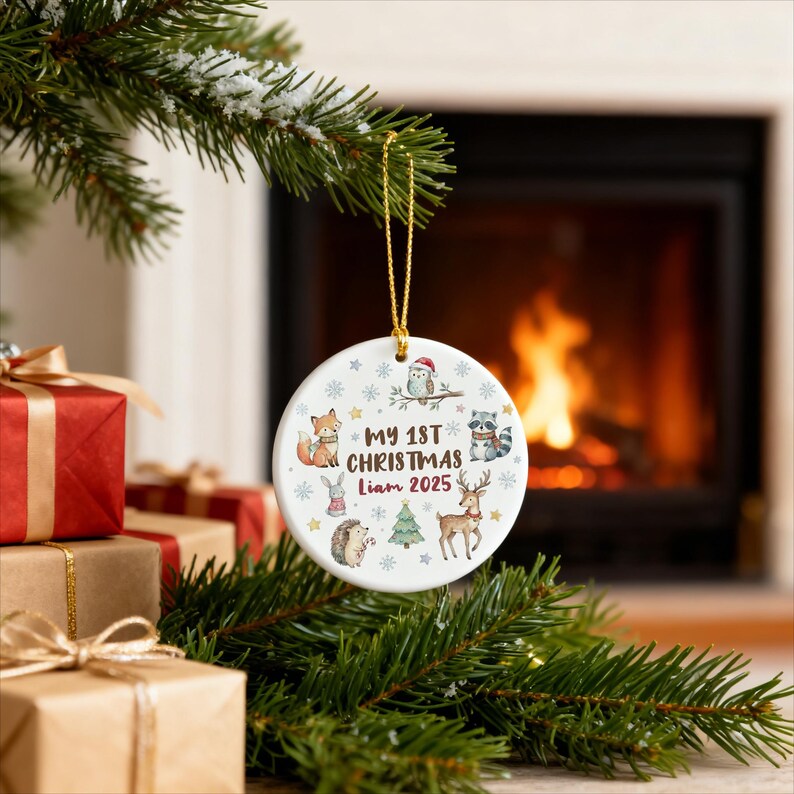 May include: A white ceramic Christmas ornament with a gold string, featuring woodland animal illustrations and the text "MY 1ST CHRISTMAS Liam 2025". The ornament is decorated with snowflakes and a Christmas tree.