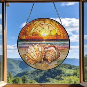 May include: Round stained glass window art depicting a beach scene with two seashells on the sand. The sunset is in shades of orange, yellow, and red. The window is hanging from a chain.