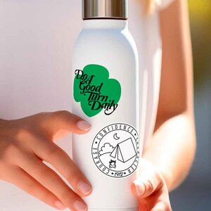 K&ouml;nnte beinhalten: Eine wei&szlig;e Wasserflasche aus Edelstahl mit einem gr&uuml;nen Kleeblatt-Design und dem Text "Do a Good Turn Daily". Die Flasche hat auch einen schwarzen und wei&szlig;en runden Aufkleber mit dem Text "Mut, Selbstvertrauen, Charakter, 1912" und einer Campingzeltgrafik.