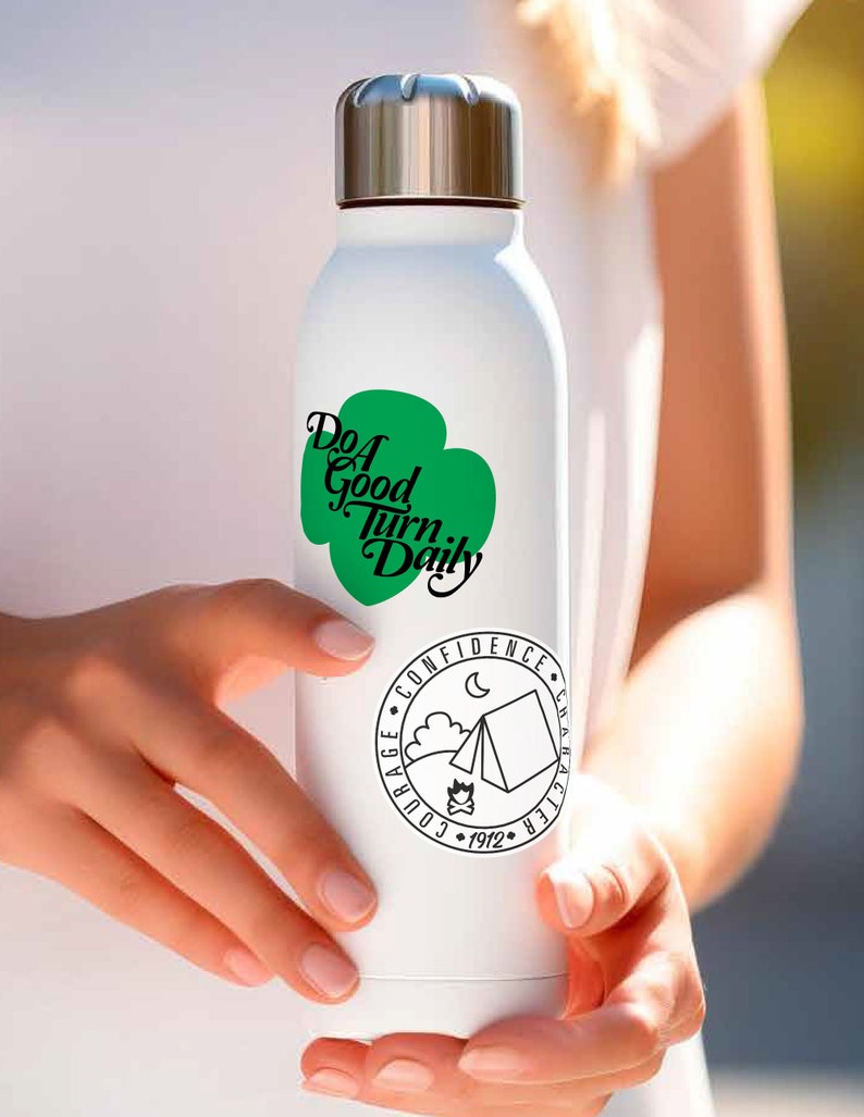 K&ouml;nnte beinhalten: Eine wei&szlig;e Wasserflasche aus Edelstahl mit einem gr&uuml;nen Kleeblatt-Design und dem Text "Do a Good Turn Daily". Die Flasche hat auch einen schwarzen und wei&szlig;en runden Aufkleber mit dem Text "Mut, Selbstvertrauen, Charakter, 1912" und einer Campingzeltgrafik.
