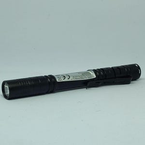 Op de afbeelding: Een zwarte zaklamp met een zakclip. De zaklamp heeft een getextureerde behuizing en een helder LED-licht aan het uiteinde. De zaklamp heeft een CE-markering en andere tekst op de zijkant.