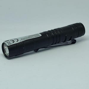 May include: A black flashlight with a silver bezel and a pocket clip. The flashlight has a ridged body for grip and a label with text. The flashlight is on a white background.