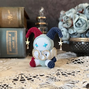 K&ouml;nnte beinhalten: Handgemachte H&auml;kelpuppe in Form eines Narren. Die Puppe hat ein wei&szlig;es Gesicht mit blau gestickten Augen und einem sternf&ouml;rmigen Anh&auml;nger. Sie hat eine rote und blaue Zipfelm&uuml;tze, einen wei&szlig;en und goldenen Kragen und sitzt auf einer Spitzefl&auml;che.