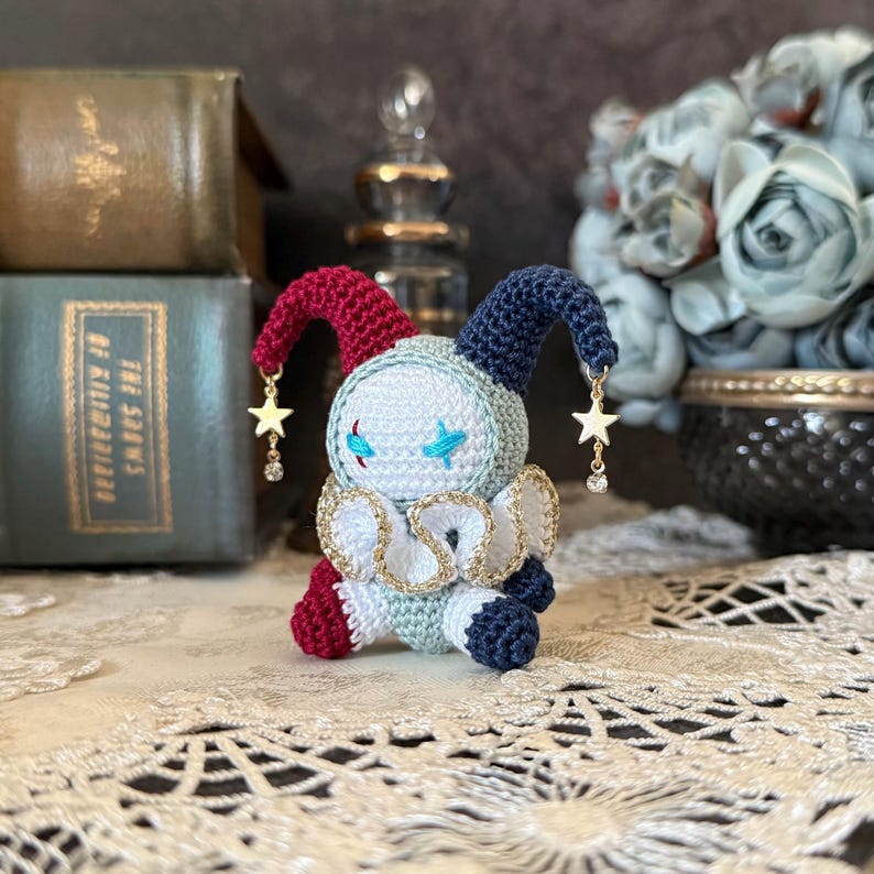 K&ouml;nnte beinhalten: Handgemachte H&auml;kelpuppe in Form eines Narren. Die Puppe hat ein wei&szlig;es Gesicht mit blau gestickten Augen und einem sternf&ouml;rmigen Anh&auml;nger. Sie hat eine rote und blaue Zipfelm&uuml;tze, einen wei&szlig;en und goldenen Kragen und sitzt auf einer Spitzefl&auml;che.