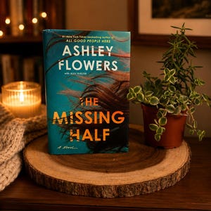 May include: A hardcover book titled "The Missing Half" by Ashley Flowers is displayed on a wooden slice. The book has a teal cover with the author's name and title in white and orange. A small potted plant and a lit candle are also visible.
