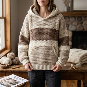 Op de afbeelding: Een gehaakte hoodie met lange mouwen en een kleurblokontwerp in crème, beige en bruin. Het kledingstuk is voorzien van een capuchon, een kangoeroezak aan de voorzijde, geribbelde boorden en een geribbelde zoom met verticale kabelstructuur.