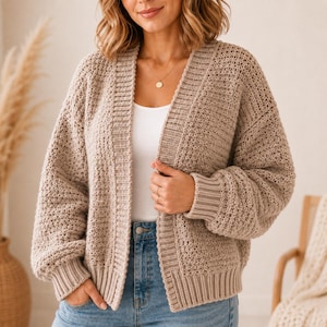 May include: A beige open-front knit cardigan with a textured stitch pattern, long sleeves, and ribbed cuffs and hem. The garment is worn over a plain white top and blue denim jeans, featuring a simple gold pendant necklace on a thin chain.