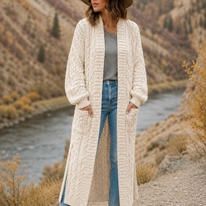 Può includere: Un lungo cardigan in maglia a trecce color crema con apertura frontale, maniche lunghe con polsini a coste e due grandi tasche applicate. Il capo è indossato sopra una t-shirt grigia e jeans blu, caratterizzato da un motivo a maglia testurizzato.