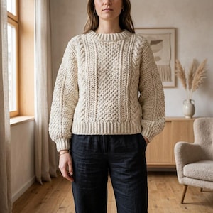 May include: A cream-colored long-sleeve sweater featuring a textured cable knit pattern, a ribbed crew neckline, and ribbed cuffs. The sweater has a thick, chunky knit construction with vertical cable details running down the front and sleeves.