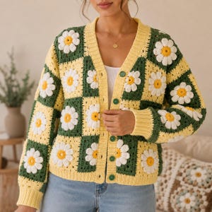 May include: A long-sleeve crochet cardigan featuring a checkerboard pattern of dark green and light yellow squares. Each square contains a white daisy motif with a yellow center. The cardigan has a V-neckline and a front button closure with green buttons.