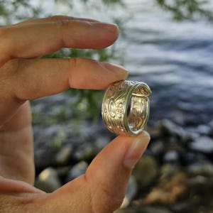 Könnte beinhalten: Ein breiter silberfarbener Ring mit einem detaillierten, geprägten Blumen- und Blattmuster auf der gesamten Außenfläche. Das Band hat eine polierte metallische Oberfläche und ein leicht offenes, gewickeltes Design mit Gravuren.
