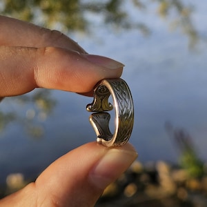 Könnte beinhalten: Ein silberfarbener Metallring mit einem komplizierten, sich wiederholenden blattartigen oder geflochtenen Muster, das in das äußere Band eingraviert ist. Der Ring hat ein offenes Design mit zwei ausgestellten, dekorativen Enden.
