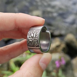 Könnte beinhalten: Ein breiter Ring aus silberfarbenem Metall mit einem geprägten Blumenmuster aus zwei Rosen mit Blättern auf einem texturierten rechteckigen Feld. Das Band hat glatte, polierte Kanten und eine Öffnung an einer Seite.