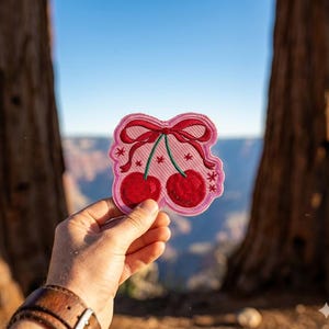May include: A pink embroidered patch featuring two red cherries with green stems and a red bow. The patch has a pink border with small star accents. The patch is held up against a scenic background.