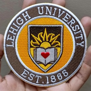 May include: Embroidered patch featuring the Lehigh University seal. The patch has a brown border with the words "LEHIGH UNIVERSITY" in white. The center has a shield design with a sun, open book, and a red heart. The text "EST. 1866" is at the bottom.