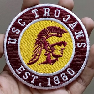 May include: A round patch with the text "USC TROJANS EST. 1880". The patch has a maroon border with white stitching. The center features a yellow circle with a maroon Trojan warrior head.