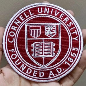 May include: A round, embroidered patch with the words "CORNELL UNIVERSITY FOUNDED A.D. 1865" in white lettering on a maroon background. The center features a shield with three distinct emblems.
