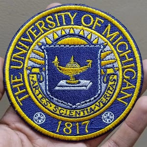 May include: Embroidered patch featuring the University of Michigan seal. The circular patch has a blue center with a gold lamp and the text "ARTES SCIENTIA VERITAS". The outer ring is yellow with the text "THE UNIVERSITY OF MICHIGAN" and the year "1817".