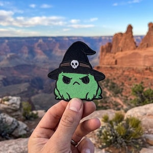 May include: Embroidered patch featuring a green creature wearing a black witch hat with a skull emblem. The creature has an angry expression. The patch is held up against a scenic background of a canyon and blue sky.