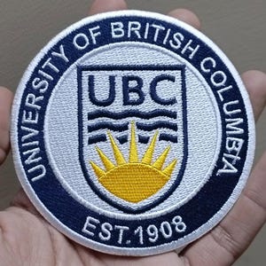 May include: Circular embroidered patch featuring the text "UNIVERSITY OF BRITISH COLUMBIA" in navy blue around the edge. The center has the letters "UBC" above a stylized sunburst design, with the text "EST. 1908" below.