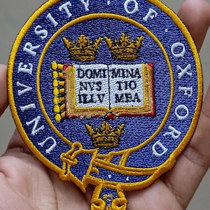 May include: Embroidered patch featuring the words "UNIVERSITY OF OXFORD" in a circular design. The patch is blue with gold trim and includes a central emblem with a book and crowns. The Latin phrase "DOMI MINA NUS TIO ILLU MEA" is visible.