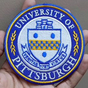 May include: Embroidered patch with the words "UNIVERSITY OF PITTSBURGH" in white on a blue background. The patch features a central emblem with a shield design, blue dots, and a gold and blue checkered pattern.