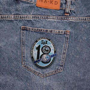 May include: Close-up of blue denim jeans with a back pocket featuring a circular patch. The patch has the text "COMMERCIAL RESUPPLY SERVICES SpX 18" with a satellite graphic in blue, black, and white.