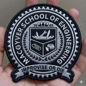 May include: A black and white embroidered patch with the text "MCGYVER SCHOOL OF ENGINEERING" encircling a central emblem. The emblem features a shield with tools and the words "IMPROVISE OR DIE" on a banner.