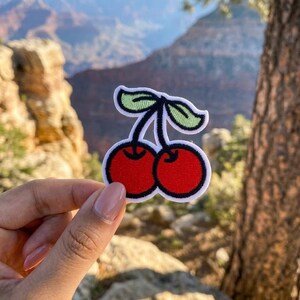 May include: Embroidered patch featuring two red cherries with green leaves and a black stem, outlined in white. The patch is held up against a scenic background of a canyon and a tree.