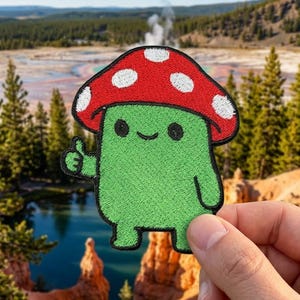 May include: Embroidered patch featuring a green mushroom character with a red cap dotted with white spots. The character is giving a thumbs-up gesture. The patch is held against a scenic background of a lake and trees.
