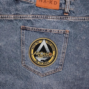 May include: Close-up of blue denim jeans with a back pocket featuring a circular patch. The patch has a gold border and the text "BACK TO THE MOON" and "ON TO MARS". The jeans have a brown leather tag that says "NA-KD".