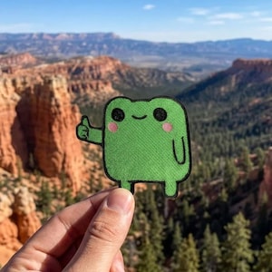 May include: A green frog patch with black eyes and pink cheeks gives a thumbs up. The patch is rectangular with rounded corners and a small U-shaped design at the bottom. The background is a scenic view of a canyon.