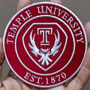 May include: A round, embroidered patch featuring the words "TEMPLE UNIVERSITY" in white lettering around the edge. The center has a red background with a white "T" and an owl design. The text "EST. 1870" is at the bottom.