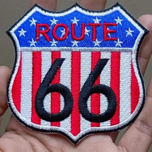 May include: Embroidered Route 66 patch in a shield shape. The top section is blue with white stars and the word "ROUTE" in red. The bottom section has red and white stripes, with the number "66" in black.