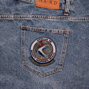 May include: Close-up of light blue denim jeans with a back pocket featuring a circular patch. The patch displays the text "APOLLO 15" and "SCOTT WORDEN IRWIN SCIENCE + J MISSION + LRY" around a graphic of a spacecraft.