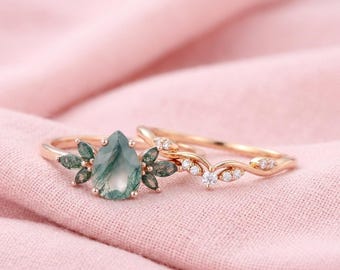 May include: Two gold rings on a pink surface. One ring features a pear-shaped, green moss agate stone with surrounding green accents. The second ring has a curved design with small, clear stones.