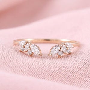 Peut inclure: Une bague en or rose avec un design à tige fendue. La bague présente des diamants taille marquise et ronds regroupés de chaque côté, créant un arrangement en forme de feuille. La bague est posée sur un fond rose.