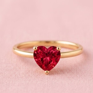 Può includere: Un anello d'oro con una gemma rossa a forma di cuore. L'anello è posto su una superficie rosa, che mette in risalto il colore vibrante della gemma. La fascia dell'anello è in oro lucido.